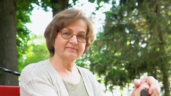 Portrait of Elderly Woman with Glasses and Cane Smiling on Bench in Public City Park