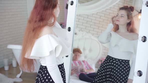 Charming Redhead Woman Trying on Outfit in Front of Mirror with Reflection of Young Man Reading Book