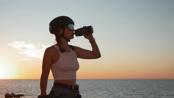 Stylish woman in top and jeans shorts wearing cycling helmet drinks water at sunset.