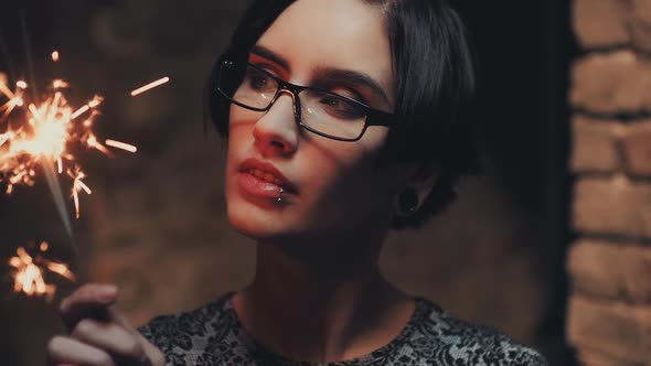 Portrait of Young Goth Woman with Short Black Hair, Wearing Glasses. He Looks at a Burning Sparkler