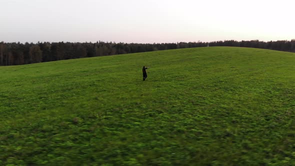 Top View of a Young Girl in a Black Trench Coat Walking Across a Green Field with Her Hands Up to