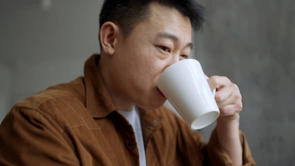 Laughing Asian man drinking coffee and watching video on tablet