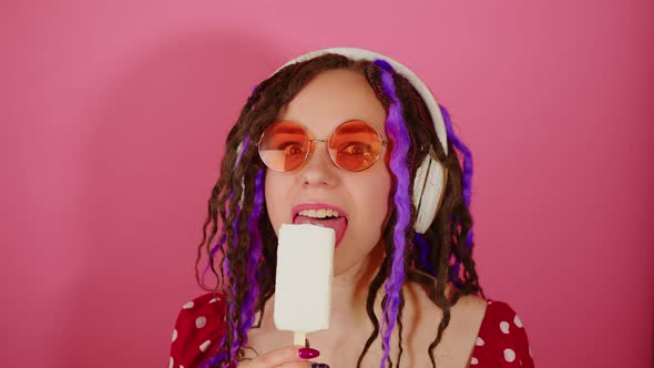 Young beautiful woman in bright sunglasses, wireless white headphones licking ice cream