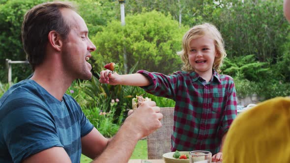 Smiling caucasian boy feeding his father a strawberry during family meal in garden