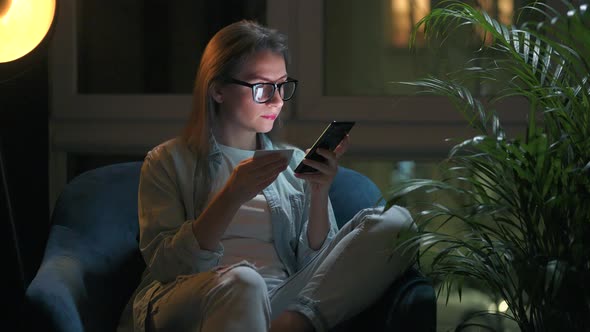 Woman with Glasses is Sitting in the Armchair and Using Smartphone at Night at Night