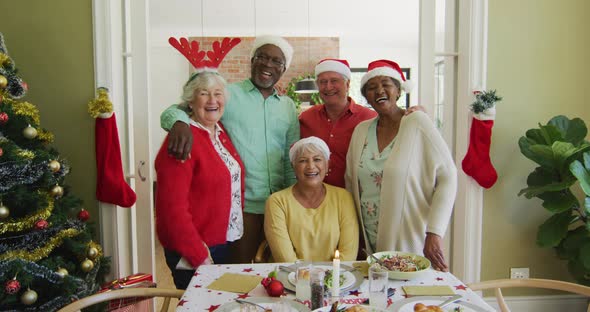 Portrait of diverse senior friends in christmas hats embracing and smiling at christmas dinner table
