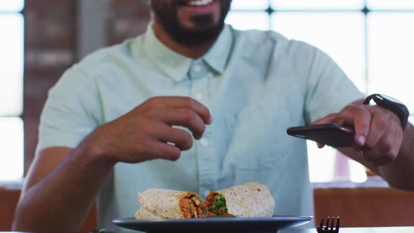 Happy mixed race man sitting in cafe taking picture of his sandwich with smartphone and smiling